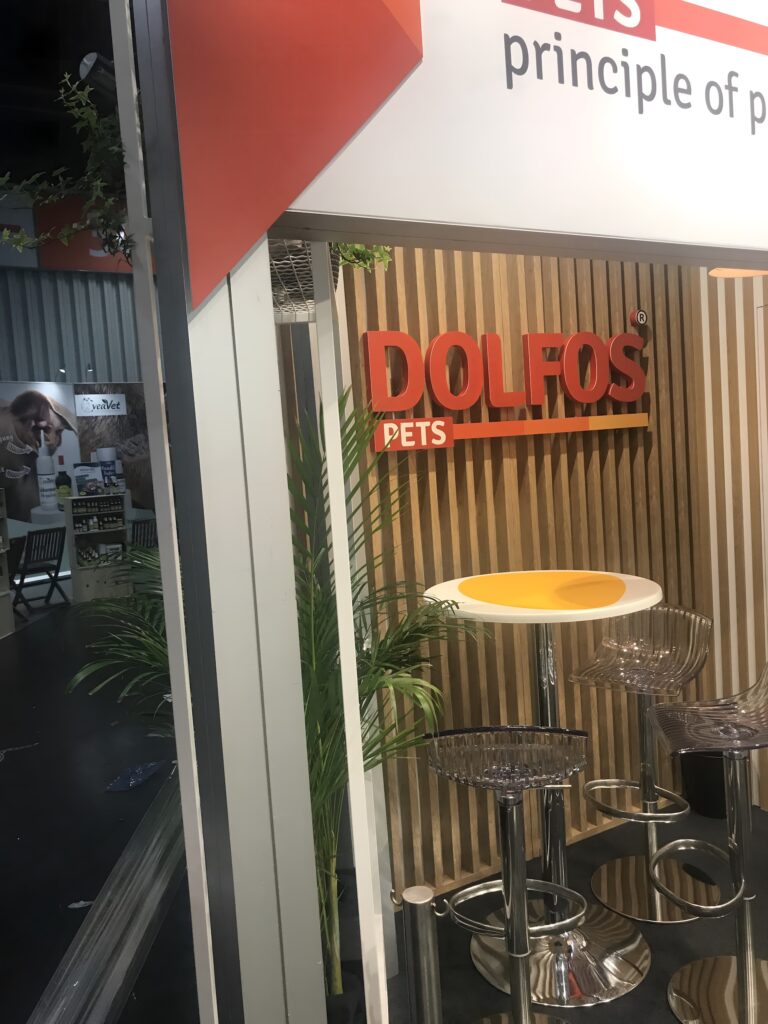 Na stoisku z drewnianą ścianą widnieje czerwony napis "DOLFOS" i zielona etykieta "PETS". Z przodu stoją dwa przezroczyste stołki barowe i mały okrągły żółty stolik. Stoisko Dolfos Pets na targach Interzoo Norymberga stoi w pobliżu rośliny. projekt i nadzór Tatjana Kobuszewska