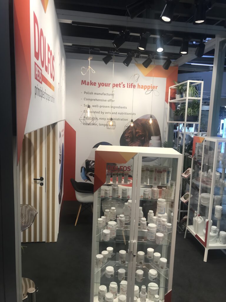 Stoisko dolfos pets na targach Interzoo Norymberga prezentuje suplementy dla zwierząt w białych butelkach na szklanych półkach. Zachęcające stoisko ma jasne tło, wygodne siedzenia, rośliny i podkreśla "Uczyń życie swojego zwierzaka szczęśliwszym". projekt i nadzór Tatjana Kobuszewska