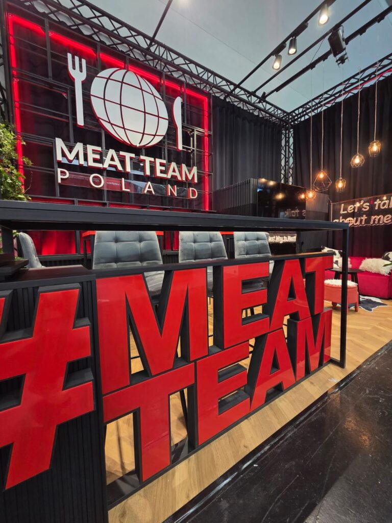 Nowoczesne stoisko z dużym czerwonym napisem MEATTEAM i przestrzennym logo Meat Team Poland podświetlone czerwonymi LED-barami-stoisko C122, Anuga 2025. Projekt Tatjany Kobuszewskiej, TRK System. Ciepłe światło i siedziska tworzą zachęcającą przestrzeń. projekt i nadzór Tatjana Kobuszewska