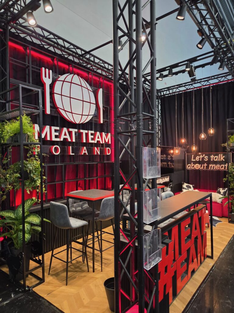 Nowoczesne stoisko wystawiennicze z oznakowaniem "MEAT TEAM POLAND" z logo kuli ziemskiej, widelca i noża na czerwono-czarnym tle. Szare stołki barowe otaczają stół, a rośliny i wiszące światła dodają dekoracji. Na pierwszym planie znajduje się duży napis MEATTEAM. projekt i nadzór Tatjana Kobuszewska