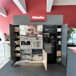 Nowoczesna ekspozycja kuchenna Miele na Interioro Design Days ICE Kraków obejmuje otwarte szafki, ekspres do kawy Miele CVA 7845, zmywarkę, piekarnik i starannie ułożoną zastawę stołową. Dobrze oświetlona aranżacja została zorganizowana na tle czerwonej ściany z białym logo Miele. Projekt Tatjany Kobuszewskiej, TRK System.