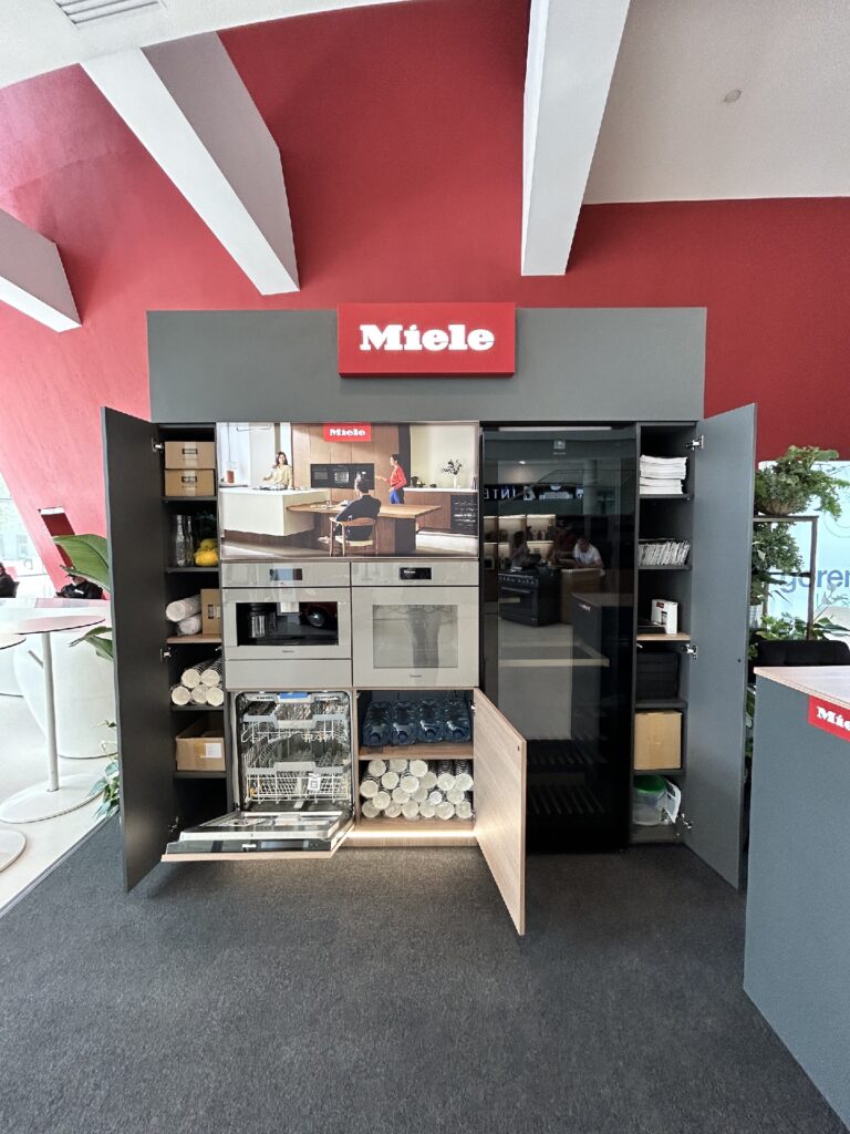 Nowoczesna ekspozycja kuchenna Miele na Interioro Design Days ICE Kraków obejmuje otwarte szafki, ekspres do kawy Miele CVA 7845, zmywarkę, piekarnik i starannie ułożoną zastawę stołową. Dobrze oświetlona aranżacja została zorganizowana na tle czerwonej ściany z białym logo Miele. Projekt Tatjany Kobuszewskiej, TRK System.