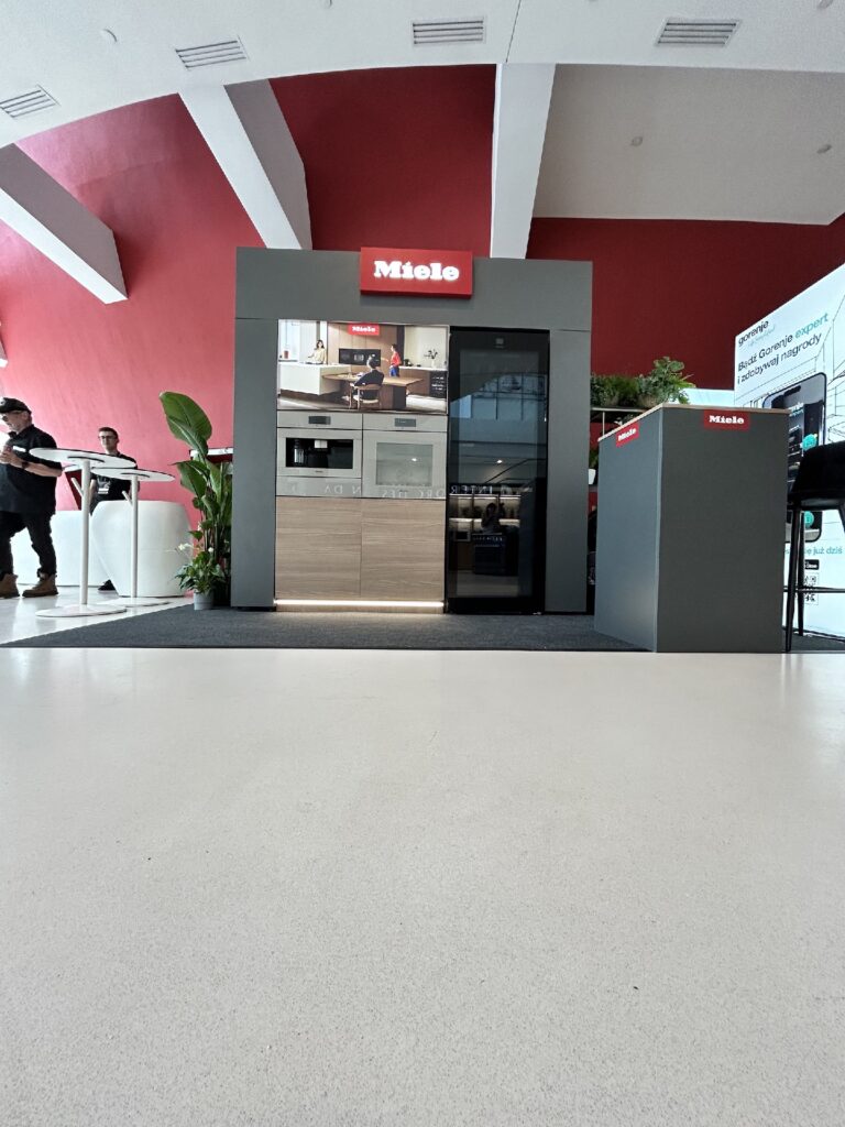 Nowoczesna ekspozycja urządzeń kuchennych Miele obejmuje lodówkę, piekarnik i kuchenkę mikrofalową Miele DGC 7865 HCX Pro w drewnianych i czarnych szafkach. Ludzie stoją w pobliżu w przestronnym, jasnym salonie wystawowym z odważną czerwoną ścianą akcentującą i roślinami do dekoracji. Projekt Tatjany Kobuszewskiej, TRK System.