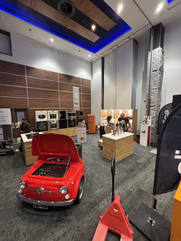 Stoisko Smeg na szkoleniu z urządzeń kuchennych, wyeksponowanych wzdłuż wyłożonych drewnem ścian. Na pierwszym planie czerwony fiat 500 lodówka/kuchenka
imitacja samochódu z otwartą maską odsłania kuchenkę - wyjątkową atrakcję w szkoleniu Smeg w hotelu Nosalowy Dwór w Zakopanem. . Projekt Tatjany Kobuszewskiej, TRK System.