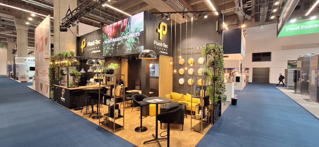 Nowoczesne stoisko targowe Plant-Tec w czarno-żółtej kolorystyce z brandingiem "Plant Based Products", stylowymi siedzeniami, wiszącymi lampami, zielenią i dekoracyjnymi półkami. Stoisko wyróżnia się w przestronnej, dobrze oświetlonej hali wystawowej z niebieskimi chodnikami wyłożonymi wykładziną. projekt i nadzór Tatjana Kobuszewska