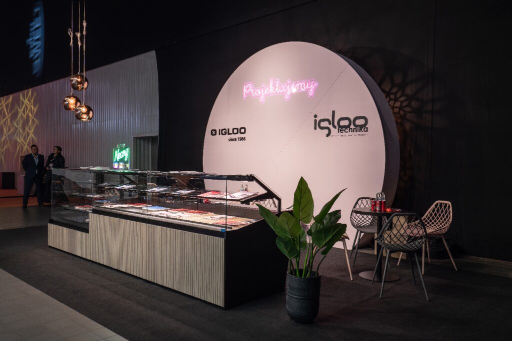 stoisko iglootechnika w stylu minimal-sweet. targi Expo Sweet w hali Expo XXI. W nowoczesnej witrynie z deserami pod szkłem wyeksponowano wypieki. Za nią widać okrągłą białą ścianę z napisem w neonowo-różowym kolorze i logo „Igloo”. Po prawej stronie, obok dużej rośliny doniczkowej, stoją dwa krzesła i stolik. Z sufitu zwisają ozdobne lampki; w tle stoją dwie osoby. projekt i nadzór Tatjana Kobuszewska