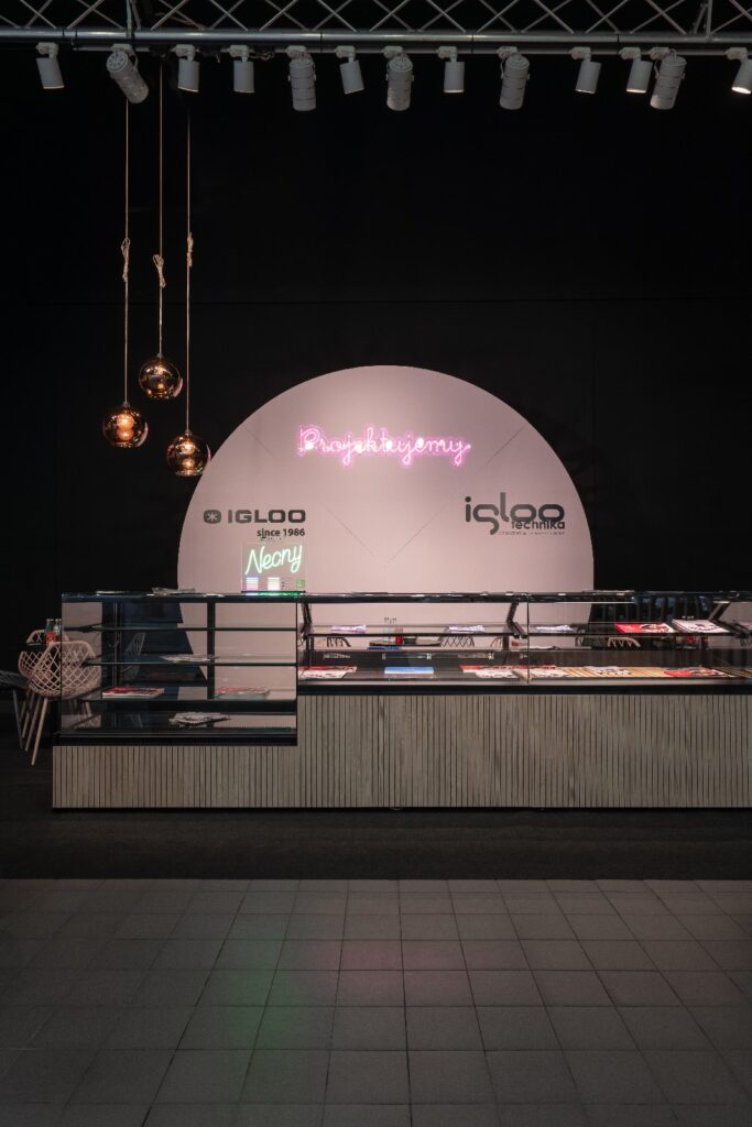 Nowoczesne stoisko „Iglootechnika” w stylu minimal-sweet, z pustymi szklanymi gablotami, brązowymi lampami wiszącymi oraz neonowymi napisami „IGLOO” i „Neony” — idealne na targi Expo Sweet w hali Expo XXI. projekt i nadzór Tatjana Kobuszewska