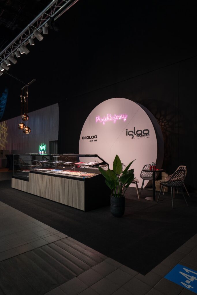 Nowoczesne stoisko wystawowe na targach Expo Sweet w hali Expo XXI to stoisko firmy Iglootechnika utrzymane w stylu „minimal-sweet”, z zakrzywionym białym tłem, neonowo-różowym napisem „Pastrylery” i czarnym logo „Igloo”, drewnianymi ladami, wypiekami oraz stylowymi akcentami. projekt i nadzór Tatjana Kobuszewska