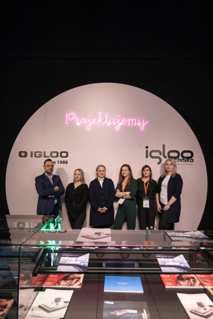 Załoga iglootechnika :bogumiła blicharska, agnieszka goryl pozuje do zdjęcia grupowego przy stoisku „Iglootechnika” w stylu minimal-sweet podczas targów Expo Sweet w hali Expo XXI. Za nimi znajduje się okrągłe tło z napisem „Projektujemy” wykonanym z różowego neonu oraz logo IGLOO; w pobliskich szklanych gablotach leżą czasopisma. projekt i nadzór Tatjana Kobuszewska
