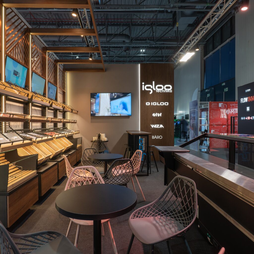 Iglootechnika Ekspozycja 360 stoiska na targach Retail Show 2024 znajdują się okrągłe czarne stoły z geometrycznymi krzesłami z drutu, drewniane półki ekspozycyjne wypełnione próbkami oraz podświetlana ściana z kasetonów z folii 3D, na której widnieją logo marek technologicznych, takich jak Iglootechnika, ATT i Niza. Na zamontowanym ekranie telewizyjnym wyświetlane są materiały. projekt i nadzór Tatjana Kobuszewska