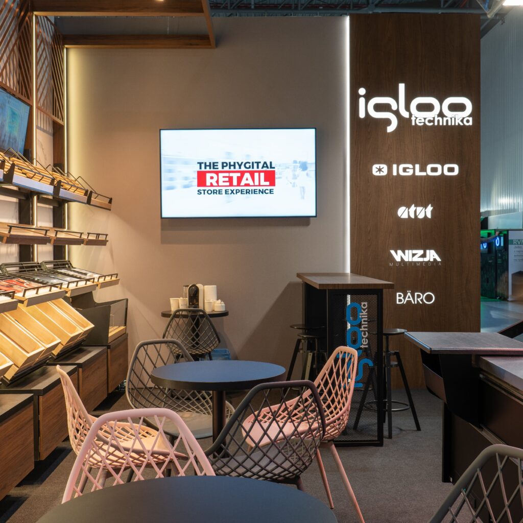 Nowoczesne stoisko targowe z geometrycznymi krzesłami i stołami, półkami na produkty po lewej stronie oraz ekranem, na którym wyświetla się napis „THE PHYGITAL RETAIL STORE EXPERIENCE”. Na prawej ścianie widnieją podświetlane logo marek — Iglootechnika, Atut, Wizja, Bäro — podczas targów Retail Show 2024. projekt i nadzór Tatjana Kobuszewska