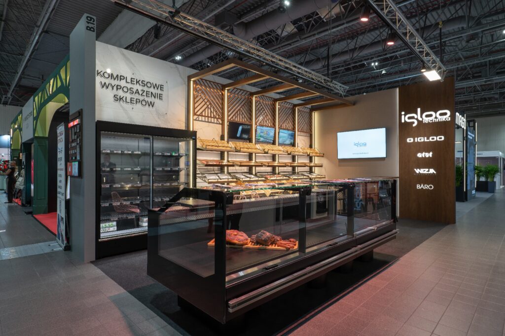 Na targach Retail Show 2024 nowoczesna ekspozycja supermarketowa obejmuje eleganckie witryny chłodnicze firmy Iglootechnika, w których prezentowane są produkty mięsne i delikatesowe. Na półkach znajdują się różnorodne produkty spożywcze, a kasetony z folii 3D oraz oświetlenie LED podkreślają napis „Kompleksowe Wyposażenie Sklepów”. projekt i nadzór Tatjana Kobuszewska