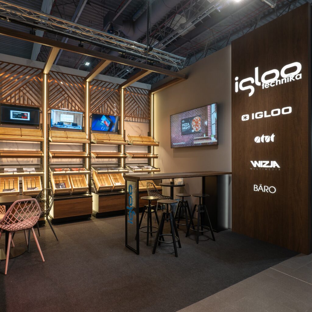 Nowoczesne stoisko targowe na targach Retail Show 2024 obejmuje próbki forniru drewnianego na stojakach ekspozycyjnych, ozdobne krzesła, podświetlane kasetony z folii 3D z logo firmy Iglootechnika oraz ekrany na tylnej ścianie pod oświetleniem sufitowym. projekt i nadzór Tatjana Kobuszewska