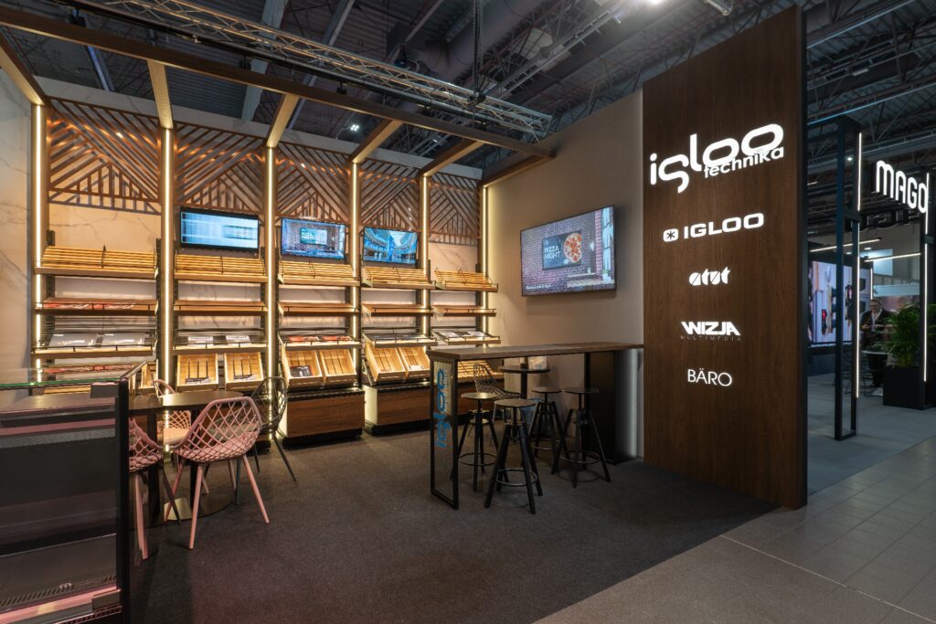 Nowoczesna ekspozycja sklepowa z drewnianymi półkami, ekranami LED oraz efektownym kasetonem z folii 3D z logo „igloo technika”. Krzesła z wikliny, wysokie stołki i stoły zachęcają gości do zanurzenia się w jasnej atmosferze targów Retail Show 2024. projekt i nadzór Tatjana Kobuszewska