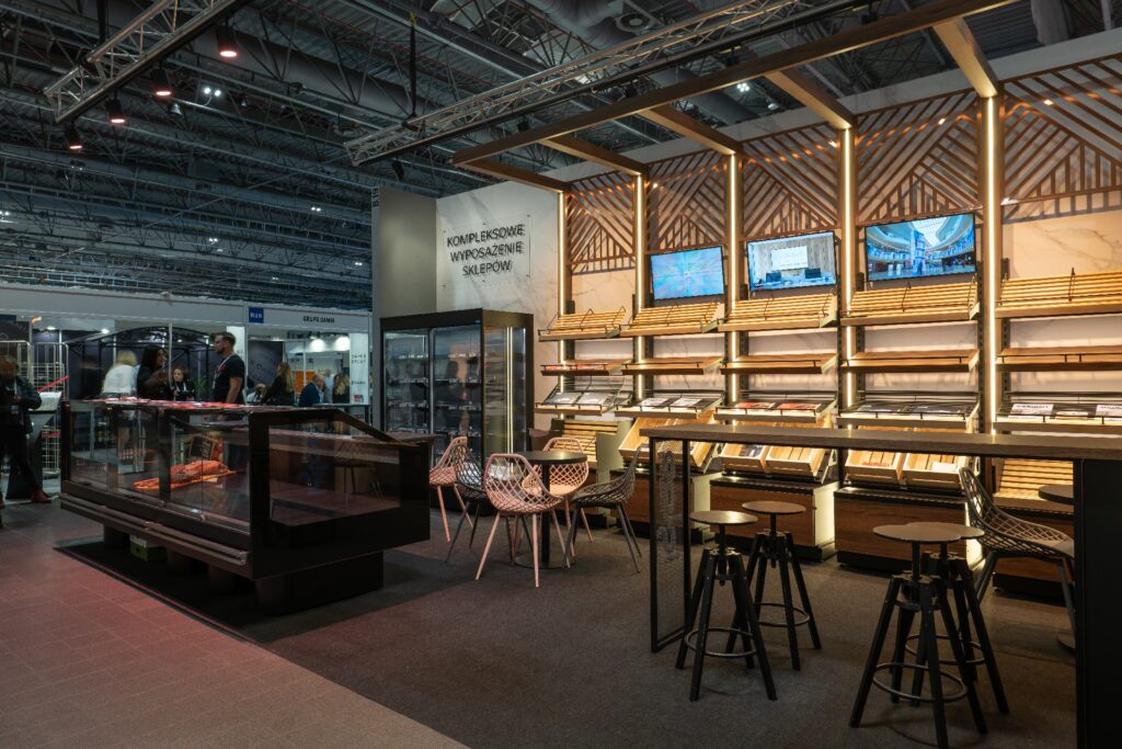 Nowoczesne stoisko targowe na targach Retail Show 2024, w którym znajdują się puste półki na pieczywo z drewnianymi akcentami, gabloty wystawowe marki Iglootechnika, trzy ekrany telewizyjne zawieszone nad nimi, niewielka strefa wypoczynkowa z okrągłymi stołami i krzesłami oraz dobrze oświetlony sufit w stylu industrialnym. projekt i nadzór Tatjana Kobuszewska