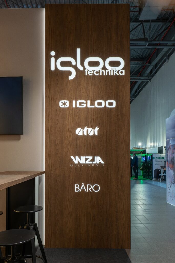 Na wysokim kasetonie wykonanym z dibondu i oklejonym specjalną folią 3D widnieją podświetlane białe logo, w tym Iglootechnika i inne, wykonane techniką kasetonów z folii 3D. Ekspozycja ta, umieszczona wewnątrz pomieszczenia w pobliżu lady i stołków, wyróżnia się na targach Retail Show 2024 pod sufitem w stylu industrialnym. projekt i nadzór Tatjana Kobuszewska