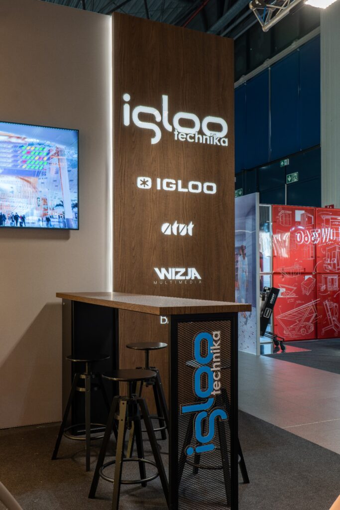 Za nowoczesnym stołem barowym na targach Retail Show 2024 stoi wysoki kaseton z folii 3D z podświetlonym logo „igloo technika”. Po lewej stronie na ekranie wyświetlane są kolorowe grafiki, a w tle widać czerwone i białe stoiska. projekt i nadzór Tatjana Kobuszewska