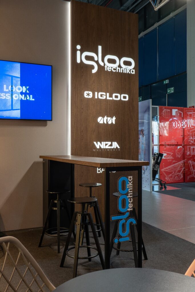Nowoczesne stoisko targowe na targach Retail Show 2024 składa się z wysokiego kasetonu z podświetlonymi logo firm Iglootechnika, IGLOO i WIZJA Multimedia. Z przodu znajduje się wysoki stół, stołki barowe oraz perforowany panel z logo „kaseton z folii 3D”. projekt i nadzór Tatjana Kobuszewska