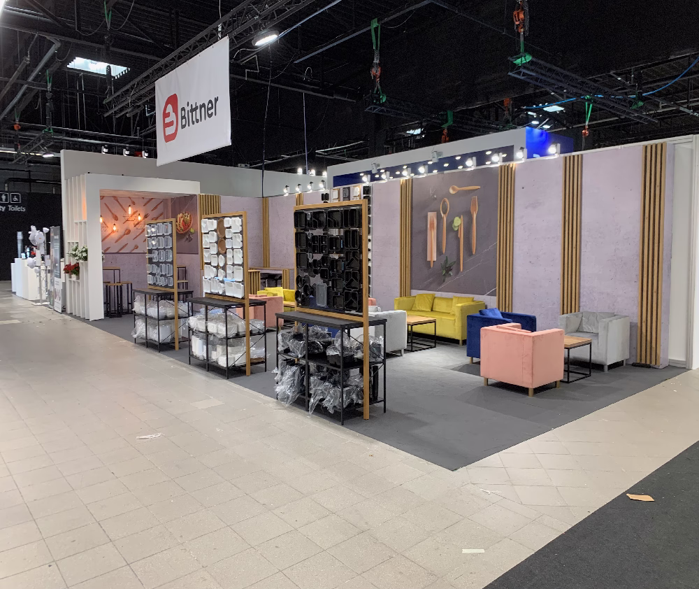 Nowoczesne stoisko targowe TRK System Warszawa z pastelowymi sofami, stojakami na poduszki i tekstylia oraz oprawionymi dziełami sztuki na liliowych ścianach. Drewniane listwy, oświetlenie górne i znak Bittner uzupełniają tę stylową aranżację stoiska targowego Warszawa na podłodze wyłożonej kafelkami. projekt i nadzór Tatjana Kobuszewska