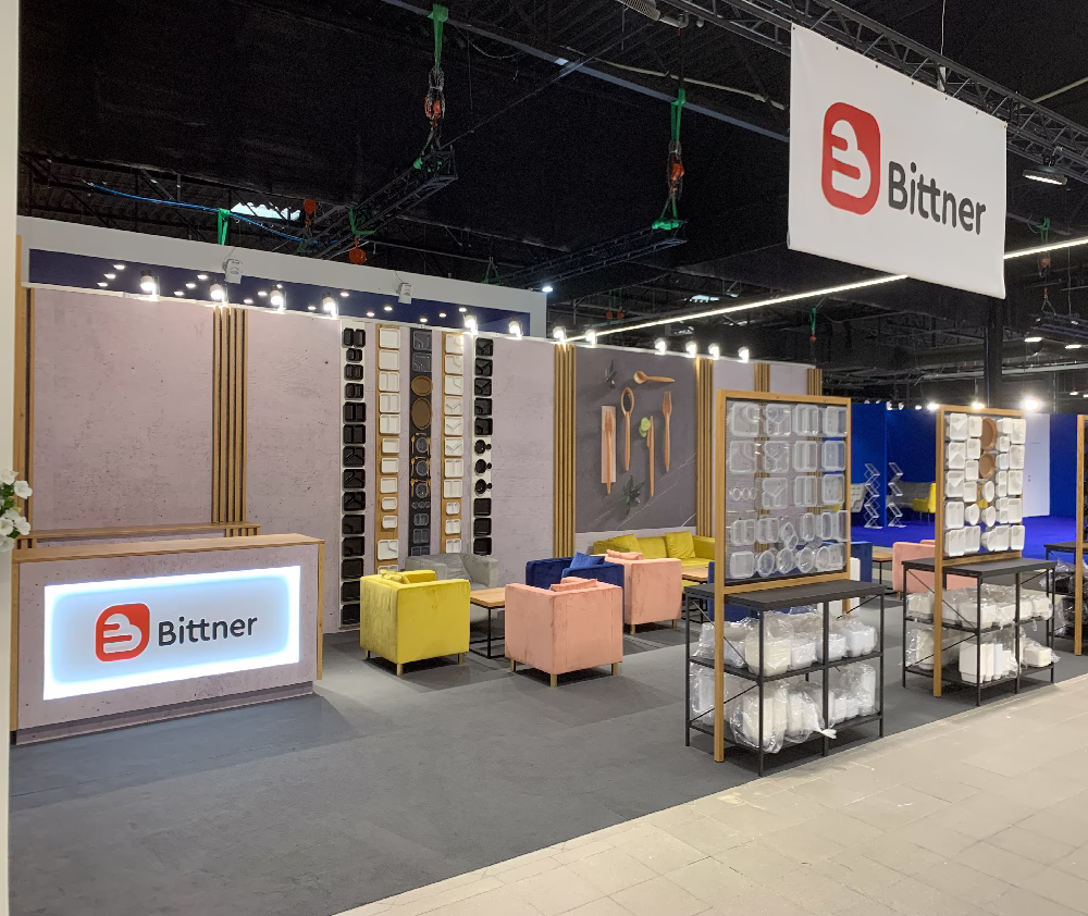 Nowoczesne stoisko wystawiennicze dla Bittner by TRK System Warszawa to podświetlana recepcja, pastelowe krzesła, ścianki ekspozycyjne z próbkami produktów oraz półki z zapakowanymi towarami - prezentacja eksperta budowa stoisk targowych Warszawa w dobrze zorganizowanym otoczeniu. projekt i nadzór Tatjana Kobuszewska