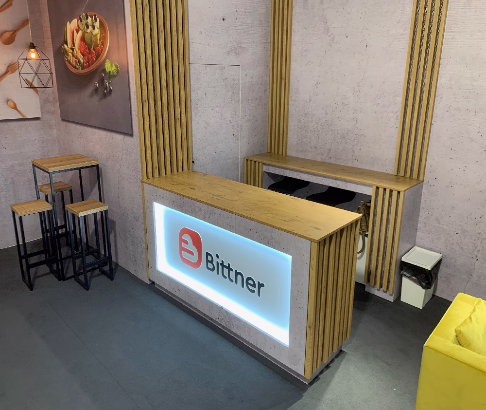 Nowoczesna recepcja lub bar zaprojektowany przez pracownię projektową stoisk, z ladą z jasnego drewna z podświetlanym logo "Bittner", wysokimi czarnymi stołkami, sztuką ścienną inspirowaną jedzeniem, akcentami z listew drewnianych, żółtym krzesłem i koszem na śmieci po prawej stronie. projekt i nadzór Tatjana Kobuszewska
