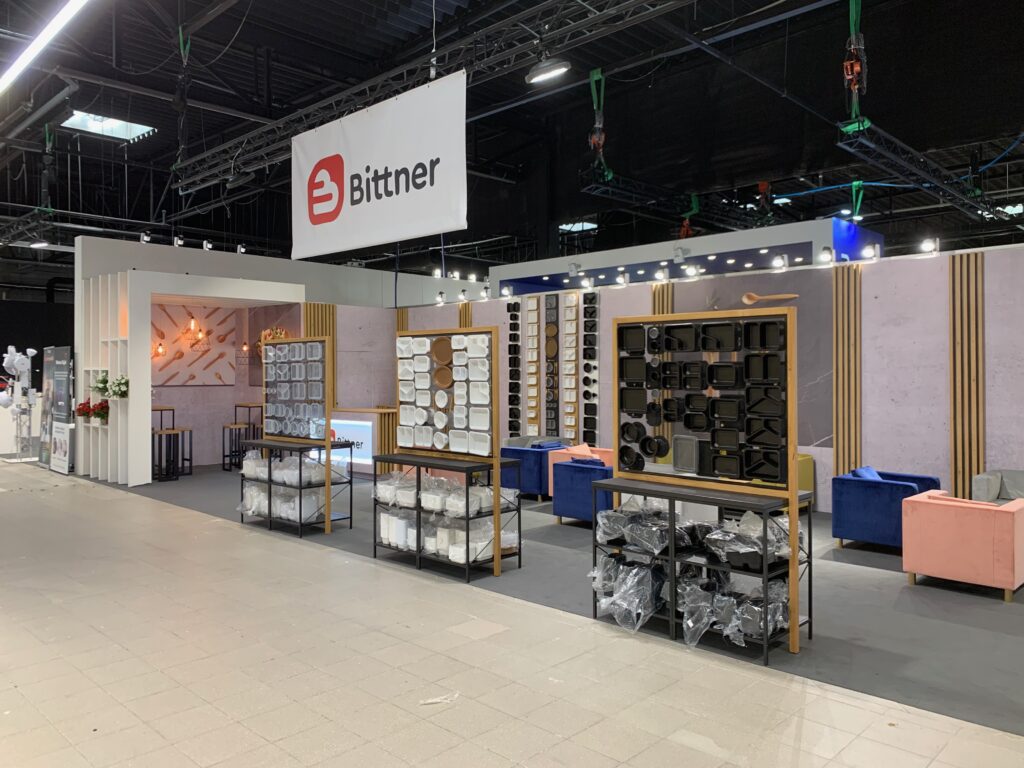 Nowoczesne stoisko targowe dla firmy Bittner, zaprojektowane przez wiodącą pracownię projektową stoisk, obejmuje eleganckie półki z uporządkowanymi przyborami kuchennymi, niebieskie i różowe siedzenia, dekoracyjne oświetlenie i widoczny napis Bittner nad głową. projekt i nadzór Tatjana Kobuszewska