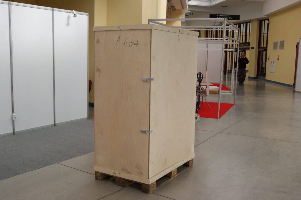 Duża drewniana skrzynia transportowa zawierająca w sobie kompletne stoisko DIY stoi na palecie w przestronnym korytarzu z betonową podłogą. W tle widoczne są stoiska wystawowe i czerwony dywan, a w oddali przechadza się człowiek. projekt i nadzór Tatjana Kobuszewska
