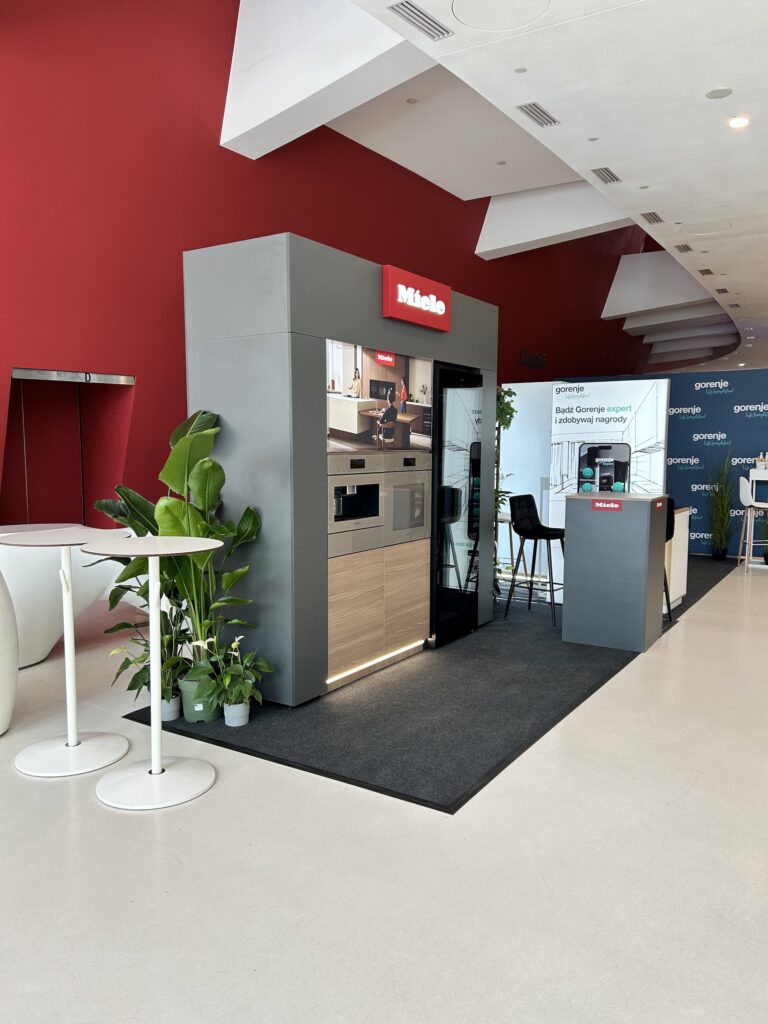 mikro stoisko Miele zawiera wbudowane ekspresy do kawy, lodówkę, zmywarkę, szufladę próżniową i. W pobliżu znajdują się wysokie białe stoły i taborety, firmy TRK System Warszawa i duża czerwona ściana w tym stylowym wnętrzu. projekt i nadzór Tatjana Kobuszewska
