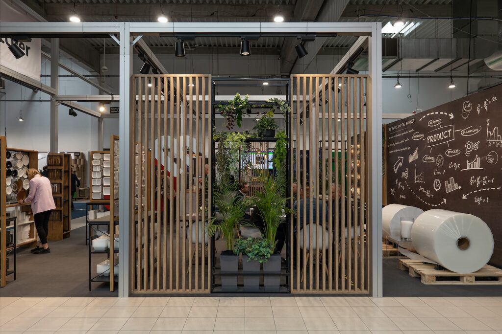 Nowoczesne industrialne wnętrze z drewnianymi ściankami działowymi, dużymi roślinami doniczkowymi i kredową ścianą ze szkicami. W pobliżu półek stoi osoba w różowej koszuli, która wydaje się być stoiskiem targowym Bittner dla Warsaw Pack. Przestrzeń sprawia wrażenie zorganizowanej i kreatywnej. projekt i nadzór Tatjana Kobuszewska