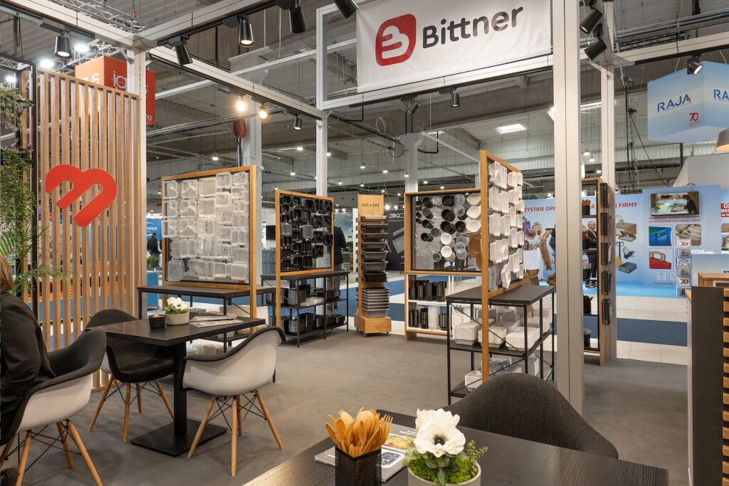 Nowoczesne stoisko targowe dla firmy Bittner na Warsaw Pack, obejmujące strefę wypoczynkową z czarno-białymi krzesłami wokół ciemnych stołów, ekspozycje produktów ze szpulami i próbkami, drewniane akcenty i banery w dobrze oświetlonej hali wystawowej. projekt i nadzór Tatjana Kobuszewska