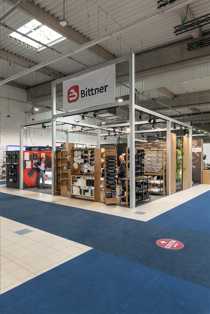 W przestronnej, dobrze oświetlonej hali centrum wystawowego Ptak Warsaw Expo ustawiono nowoczesne stoisko wystawowe firmy Bittner. Na stoisku znajdują się drewniane i metalowe ekspozytory z różnymi produktami z branży metalowej, półki z próbkami oraz panele informacyjne. Wewnątrz stoiska i wokół niego kręci się kilka osób. projekt i nadzór Tatjana Kobuszewska
