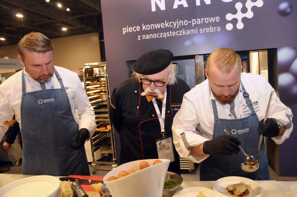 Trzech kucharzy w fartuchach stoi przy ladzie; ten pośrodku, w czarnym beretcie i okularach, gryzie drewnianą łyżkę. Pozostali skupiają się na układaniu potraw na talerzach. Przed nimi leżą przybory kuchenne i miska z jajkami. W tle widoczny jest baner z napisem w języku polskim. Ta pełna życia scena rozgrywa się na stoisku Hendi o powierzchni 250 m² podczas Kongresu Szefów Kuchni. projekt i nadzór Tatjana Kobuszewska