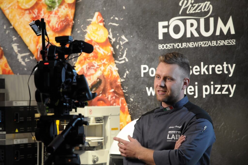Przed kamerą stoi kucharz w szarym uniformie, wskazując coś palcem i mówiąc. Za nim znajduje się ściana z dużym obrazem pizzy oraz napisem „Pizza FORUM” i „BOOST YOUR NEW PIZZA BUSINESS”. Po lewej stronie widać sprzęt kuchenny, przypominający ten, który można znaleźć na imponującym stoisku Hendi o powierzchni 250 m² na Kongresie Szefów Kuchni w hali. projekt i nadzór Tatjana Kobuszewska