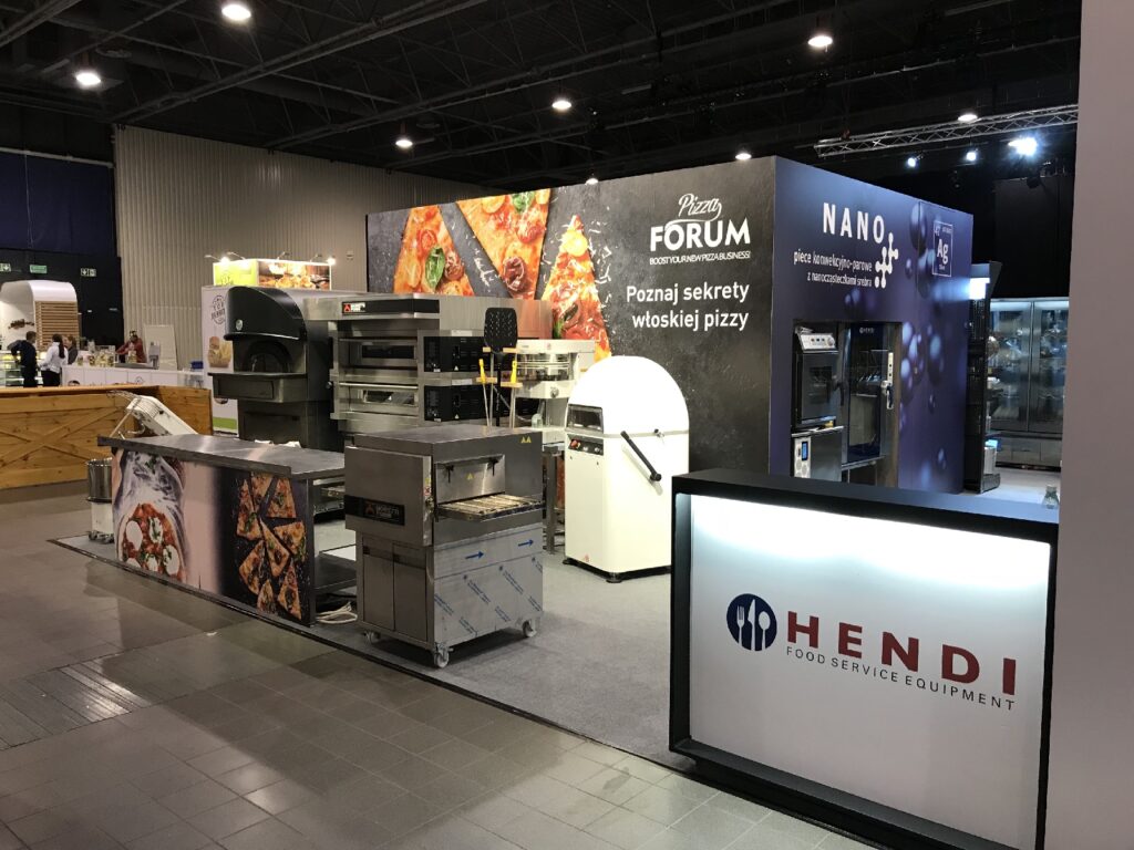 Stoisko z wyposażeniem do pizzy na targach, na którym prezentowane są piece, stoły do przygotowywania potraw oraz mikser do ciasta. Banery reklamują „Pizza Forum” oraz „Hendi Food Service Equipment”. Na ścianie w tle widnieją zdjęcia pizzy oraz tekst w języku polskim. Oświetlenie jest jasne; zdjęcie przedstawia wnętrze. projekt i nadzór Tatjana Kobuszewska