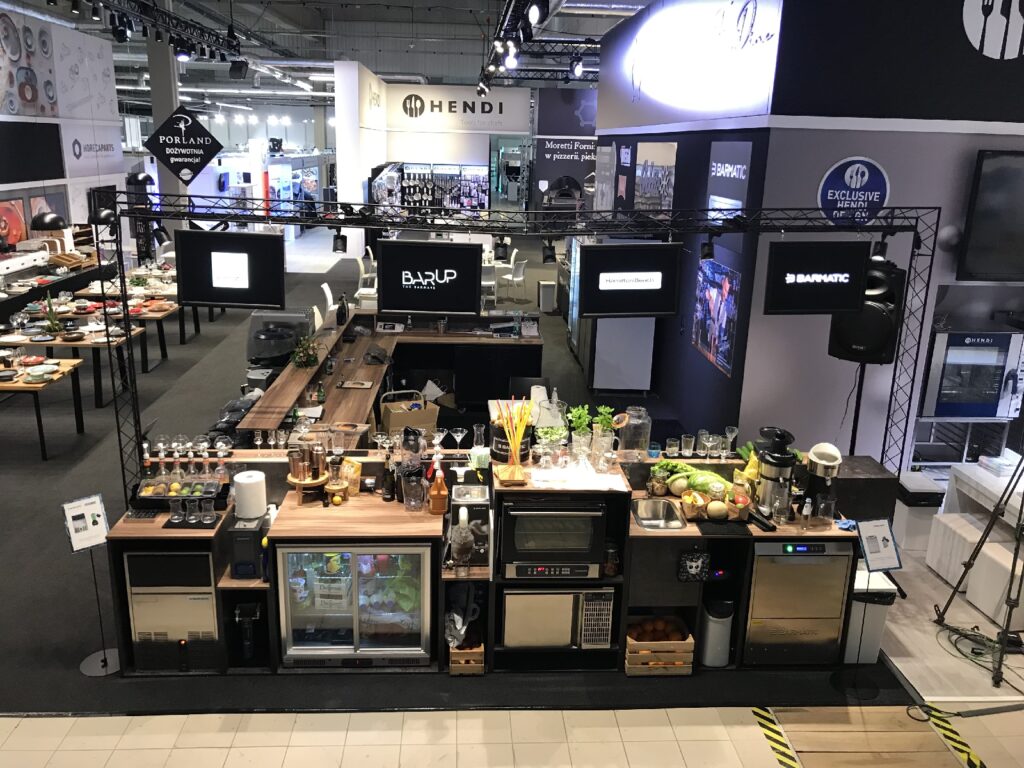 Nowoczesny blat kawiarni na targach, na którym znajdują się ekspresy do kawy, blendery, owoce, przybory kuchenne, szkło oraz świeże zioła.