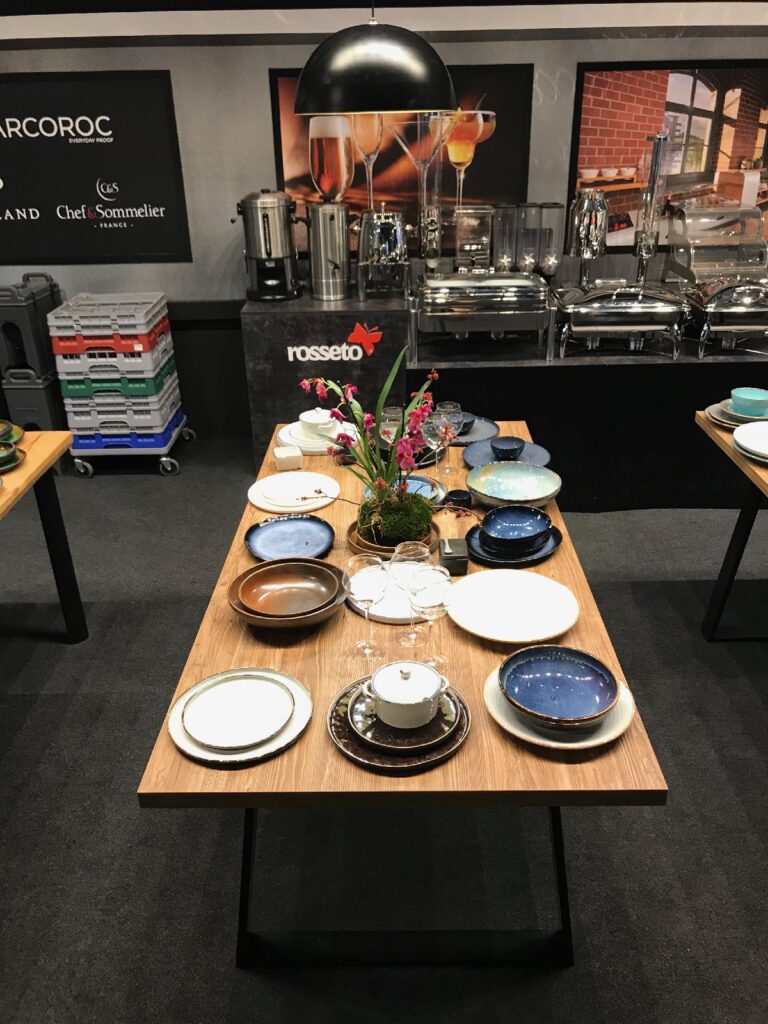 Drewniany stół nakryty różnorodnymi ceramicznymi talerzami, miskami, szklankami oraz niewielką kompozycją kwiatową.