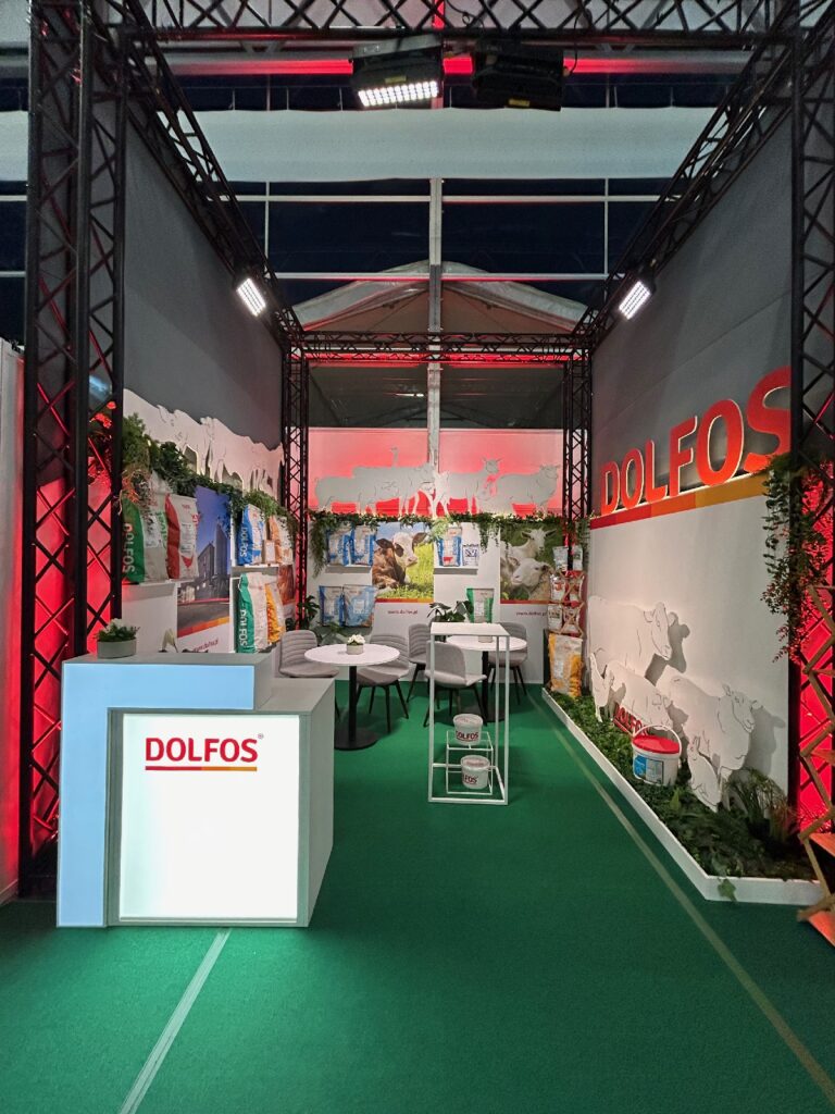 Stoisko wystawowe firmy DOLFOS na targach 2025-dolfos-agrotech-kielce-0520 obejmuje zielony dywan, worki z paszą dla zwierząt, grafiki ścienne przedstawiające zwierzęta hodowlane, białą ladę recepcyjną z logo DOLFOS, okrągłe stoły z krzesłami, rośliny oraz czerwone akcenty świetlne w całej przestrzeni. projekt i nadzór Tatjana Kobuszewska