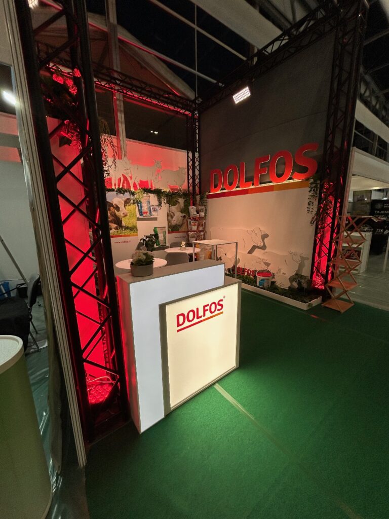 Stoisko targowe firmy DOLFOS na imprezie „2025-dolfos-agrotech-kielce-0526” składa się z białego blatu z logo DOLFOS, oświetlonego efektownym czerwonym światłem. Za ladą znajdują się grafiki przedstawiające zwierzęta, bujne rośliny oraz pojemniki z produktami. Tło i konstrukcje są podkreślone czerwono-zielonymi elementami dekoracyjnymi. projekt i nadzór Tatjana Kobuszewska