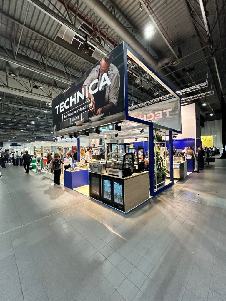 Nowoczesne stoisko „Technica” na kongresie szefów kuchni i restauratorów prezentuje duży baner przedstawiający kucharza przy pracy oraz gabloty z potrawami, napojami i sprzętem. W tle tej przestronnej, dobrze oświetlonej sali widać kilka osób zwiedzających stoisko. projekt i nadzór Tatjana Kobuszewska