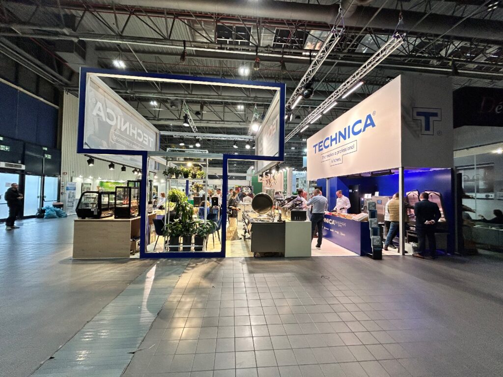 Nowoczesne stoisko targowe „Stoisko Technica” na kongresie szefów kuchni i restauratorów charakteryzuje się dużymi niebieskimi ramami, roślinami oraz ladą, przy której ludzie nawiązują kontakty pod wyrazistym napisem „TECHNICA” w przestronnej, dobrze oświetlonej hali wystawowej. projekt i nadzór Tatjana Kobuszewska