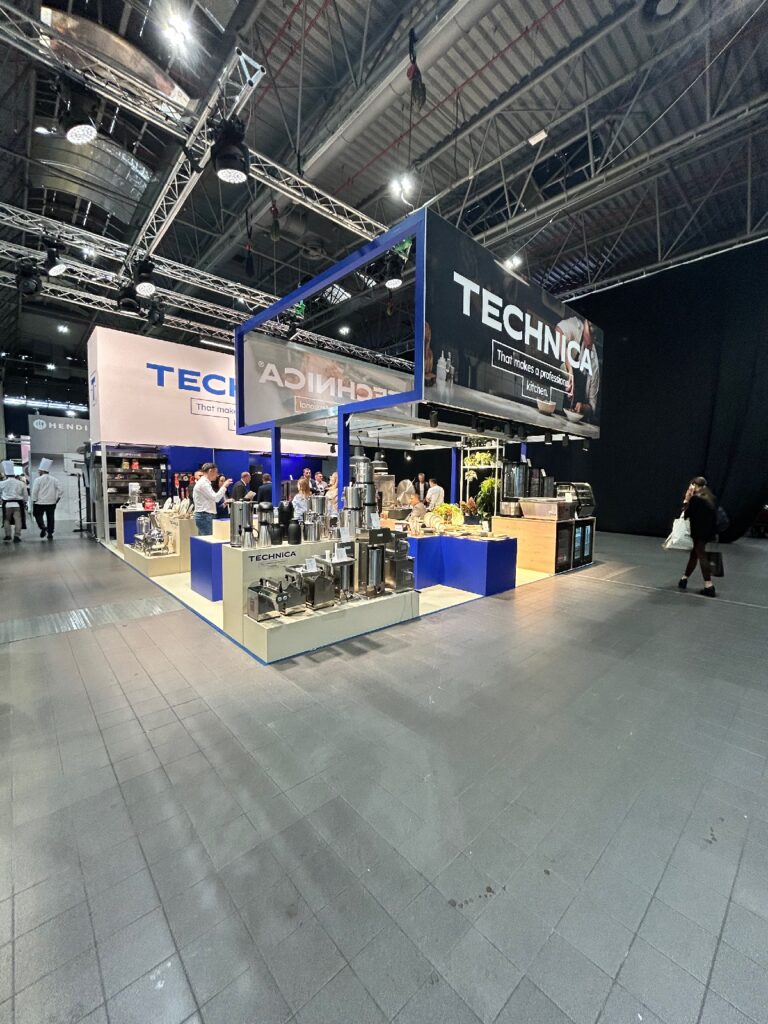 Stoisko targowe firmy TECHNICA, zrealizowane przez pracownię TRK System, prezentuje sprzęt przemysłowy na niebieskich i srebrnych ladach. Kilka osób kręci się wokół stoiska Technica, które wyróżnia się jasnym oświetleniem i dużymi banerami zawieszonymi nad nim. projekt i nadzór Tatjana Kobuszewska
