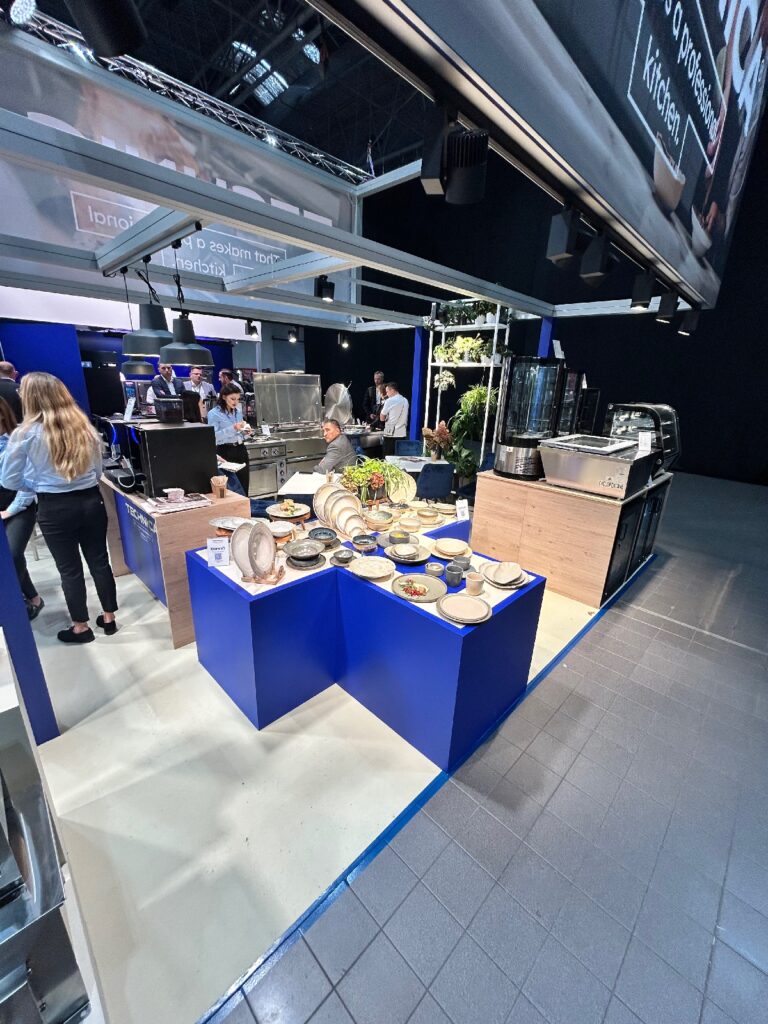 Na stoisku gastronomicznym, będącym częścią „Stoisko Technica” podczas kongresu szefów kuchni i restauratorów, na niebieskim blacie prezentowane są różne potrawy i półmiski. Goście rozmawiają z obsługą w otoczeniu sprzętu kuchennego, roślin i jasnego oświetlenia w nowoczesnej aranżacji. projekt i nadzór Tatjana Kobuszewska