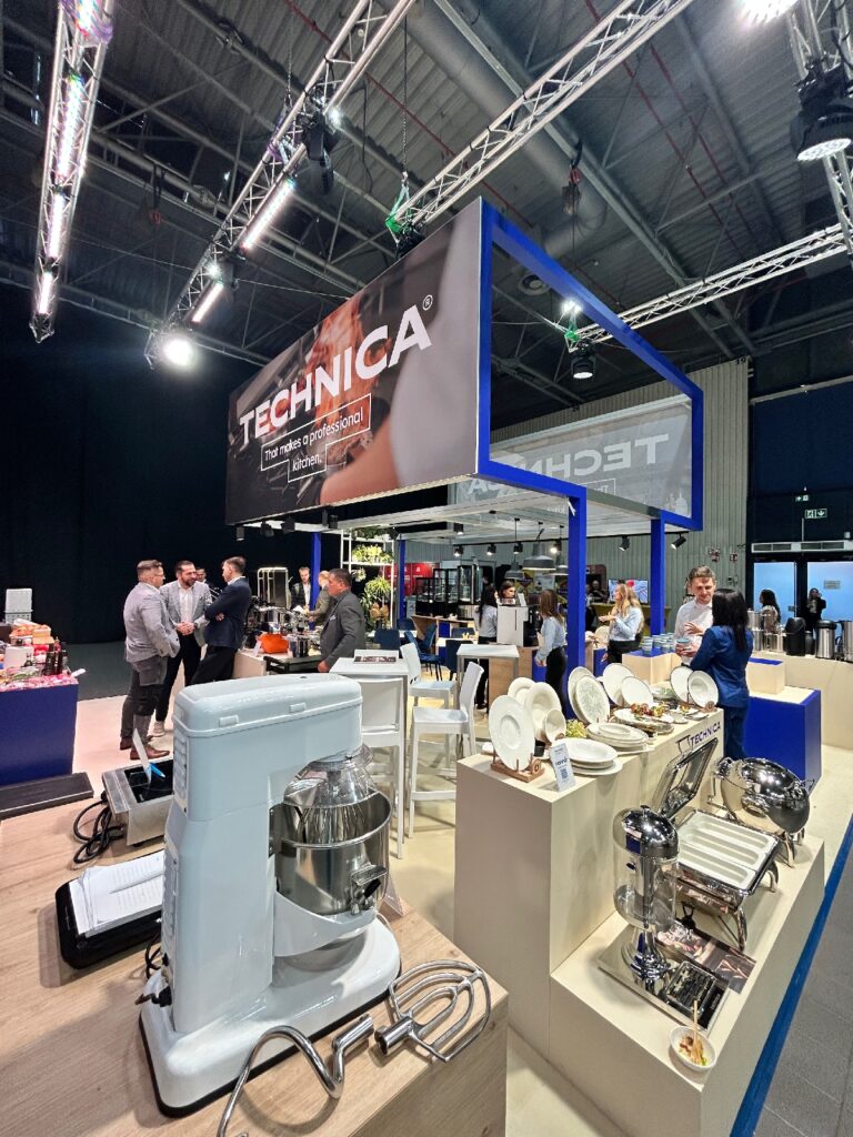 Na stoisku „Technica” podczas kongresu szefów kuchni i restauratorów prezentowane są urządzenia kuchenne i zastawa stołowa, w tym biały mikser stojący oraz srebrne podgrzewacze do potraw. Uczestnicy rozmawiają pod widocznym napisem „TECHNICA” w przestronnej, dobrze oświetlonej hali wystawowej. projekt i nadzór Tatjana Kobuszewska