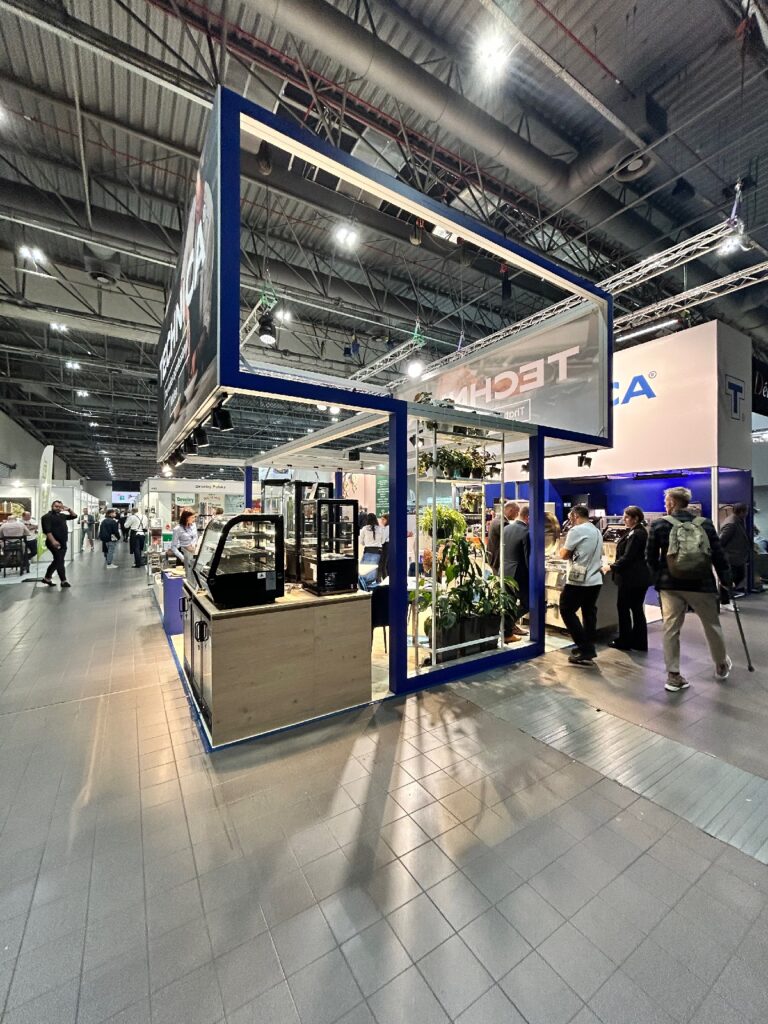 Nowoczesne stoisko targowe firmy „Ekspozycja sprzętu gastronomicznego Technica Group” charakteryzuje się niebieską konstrukcją i szklanymi gablotami. Wokół niego gromadzi się kilka osób, otoczonych jasnym oświetleniem, roślinami i innymi stoiskami wystawowymi w przestronnej hali. projekt i nadzór Tatjana Kobuszewska