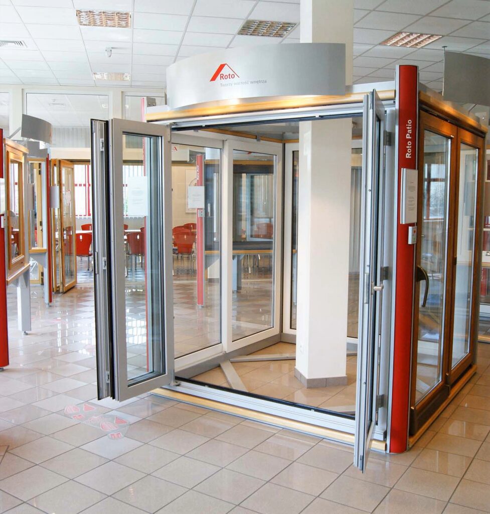 Salon wystawowy z dużymi szklanymi drzwiami tarasowymi osadzonymi w drewnianych ramach; drzwi są otwarte, ukazując swój mechanizm składania i przesuwania. W tle nowoczesnej sali szkoleniowej, prezentującej zastosowanie okuć okiennych Roto, widoczna jest podłoga wyłożona błyszczącymi płytkami, oświetlenie sufitowe, białe ściany oraz inne ekspozycje okienne. Nad ekspozycją widnieje logo Roto. projekt i nadzór Tatjana Kobuszewska