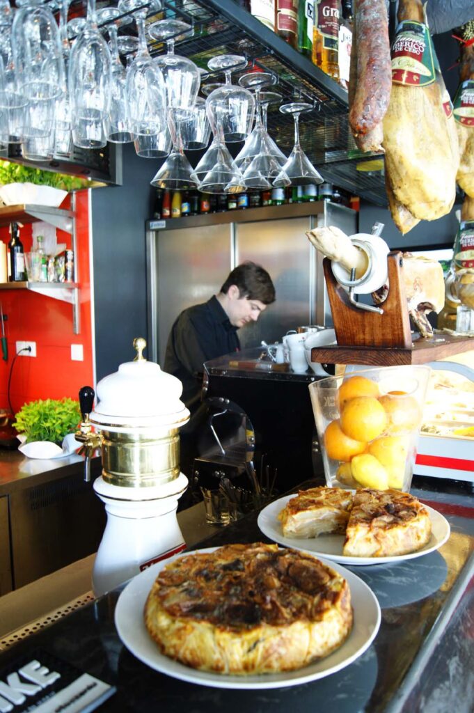 Tętniący życiem bar z tapas w Carmonie, w którym nad ladą, za którą zręcznie pracuje barman, wiszą kieliszki i wędliny. Na pierwszym planie, obok świeżych pomarańczy, zielonych warzyw liściastych i dwóch złocistobrązowych quiche’ów ułożonych na talerzach, stoi biały ekspres do kawy. Półki z butelkami wypełniają tę jasno oświetloną, kolorową przestrzeń, dodając jej przyjaznej atmosfery. projekt i nadzór Tatjana Kobuszewska
