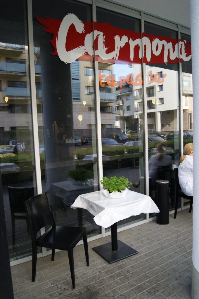 Przed restauracją Carmona Tapas Bar, której ściany są przeszklone, stoi mały stolik nakryty białym obrusem i ozdobiony zielonymi roślinami. Przy stoliku stoją dwa czarne krzesła. W pobliżu siedzą ludzie. W szybie widać odbicia samochodów i budynków. projekt i nadzór Tatjana Kobuszewska