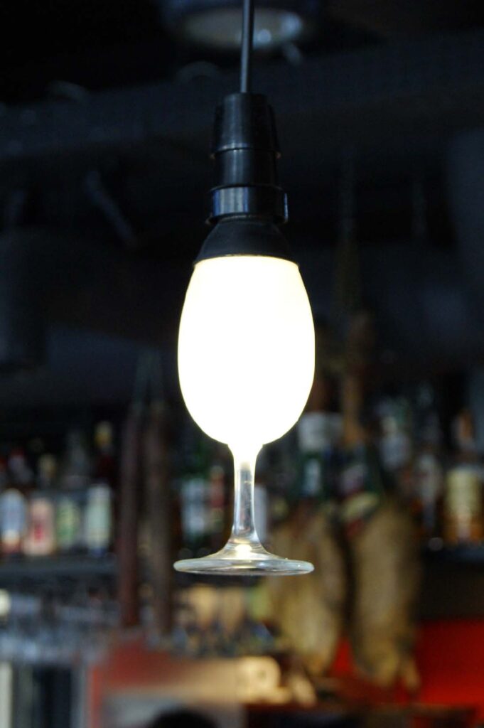W lokalu Carmona Tapas Bar Warszawa z czarnego przewodu zwisa żarówka w kształcie odwróconego kieliszka do wina. Wyraźnie widać nóżkę i podstawę kieliszka, a świecąca żarówka tworzy jego czaszę. Na rozmyłym tle widać półki z butelkami, co potęguje niepowtarzalny klimat inspirowany lampą autorstwa P. Gołębiewskiego. projekt i nadzór Tatjana Kobuszewska