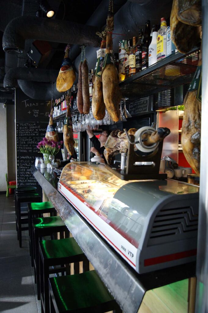 Skąpany w słońcu bar Carmona Tapas Bar Warszawa z wiszącymi wędlinami, półkami z alkoholami, chłodzoną witryną z potrawami oraz stołkami ustawionymi wzdłuż baru ozdobionego kwiatami. Wiszące kieliszki i tablice z ręcznie pisanymi menu dodają temu miejscu przytulnej atmosfery. projekt i nadzór Tatjana Kobuszewska
