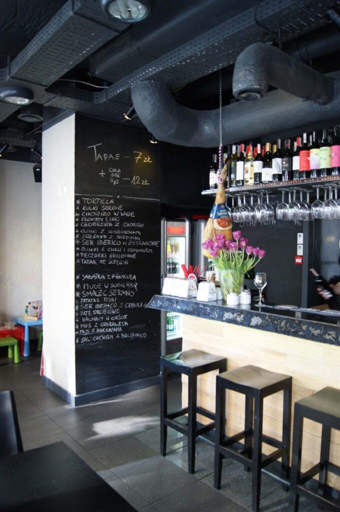 Nowoczesny bar tapas „Carmona” z wysokimi stołkami przy drewnianym blacie, menu zapisanym kredą na tablicy na ścianie, tulipanami na barze, wiszącymi kieliszkami do wina oraz butelkami wina wyeksponowanymi na półkach. W tle widać przemysłowe rury sufitowe oraz kącik zabaw dla dzieci. projekt i nadzór Tatjana Kobuszewska